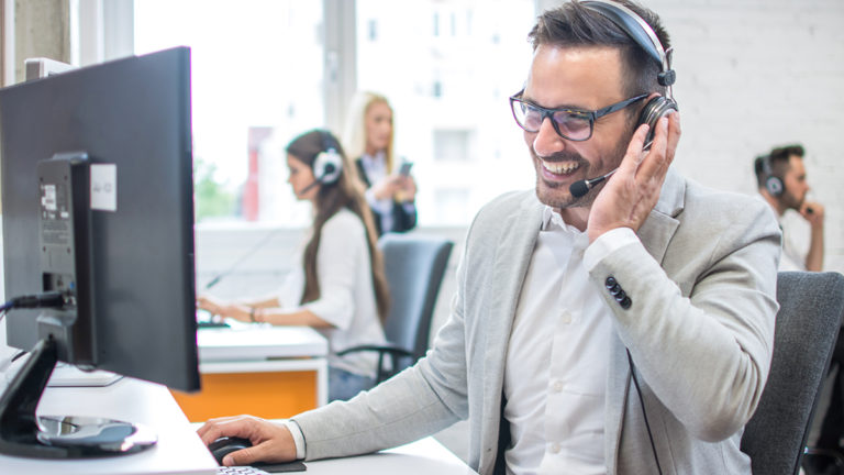 Businessman with a large smile on the phone with a voice over IP headset looking at his monitor sitting in an office environment shown blurred in the background