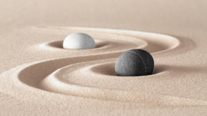 One round dark stone and one round light stone in sand with a swirl drawn between them representing the balance of yin and yang