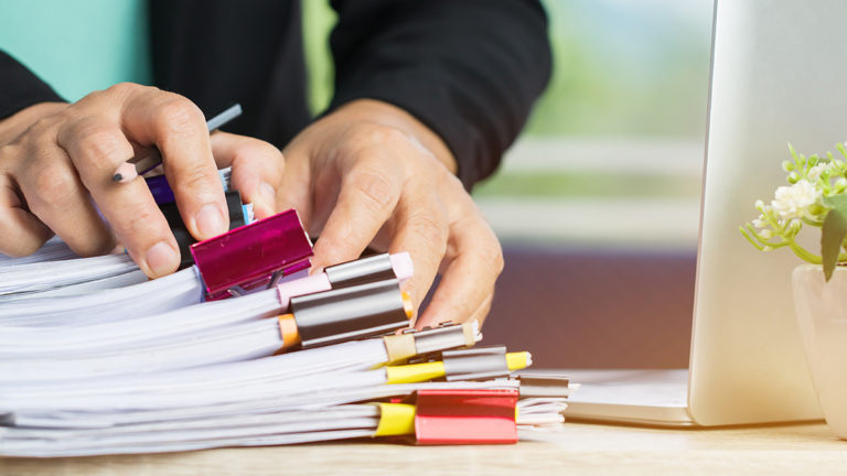 Businessman hands holding pencil for working in Stacks of paper files searching regulatory compliance documentation papers on computer desk with laptop and small plant.