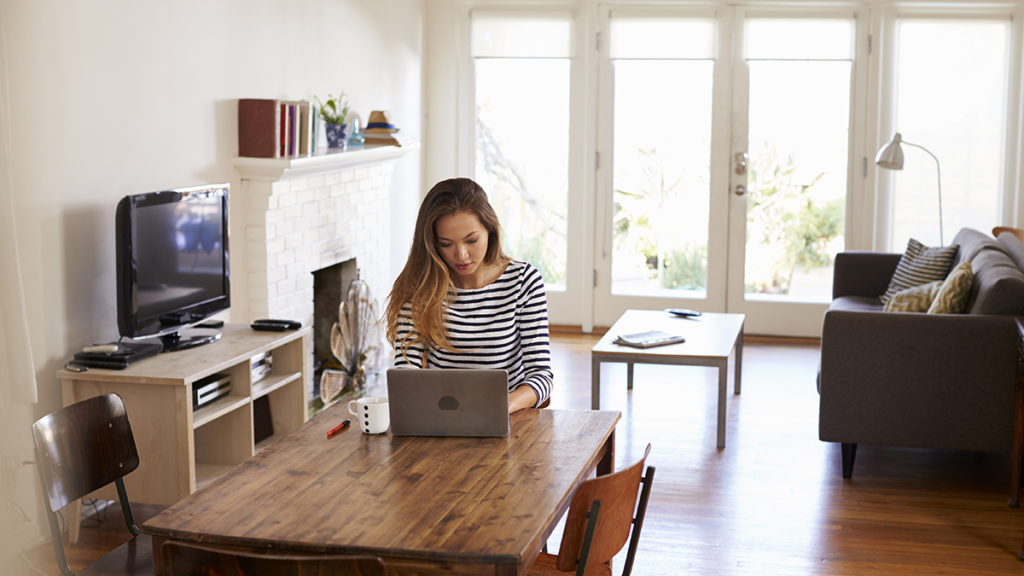 Young woman in a striped shirt working on her laptop on a wooden dining table in her home with her living room in the background.